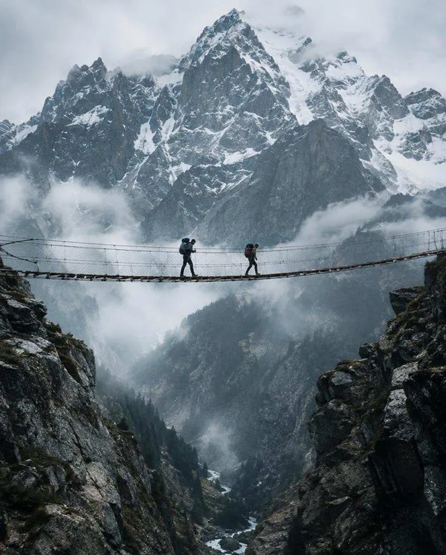Two hikers crossing a narrow suspension bridge above a misty gorge